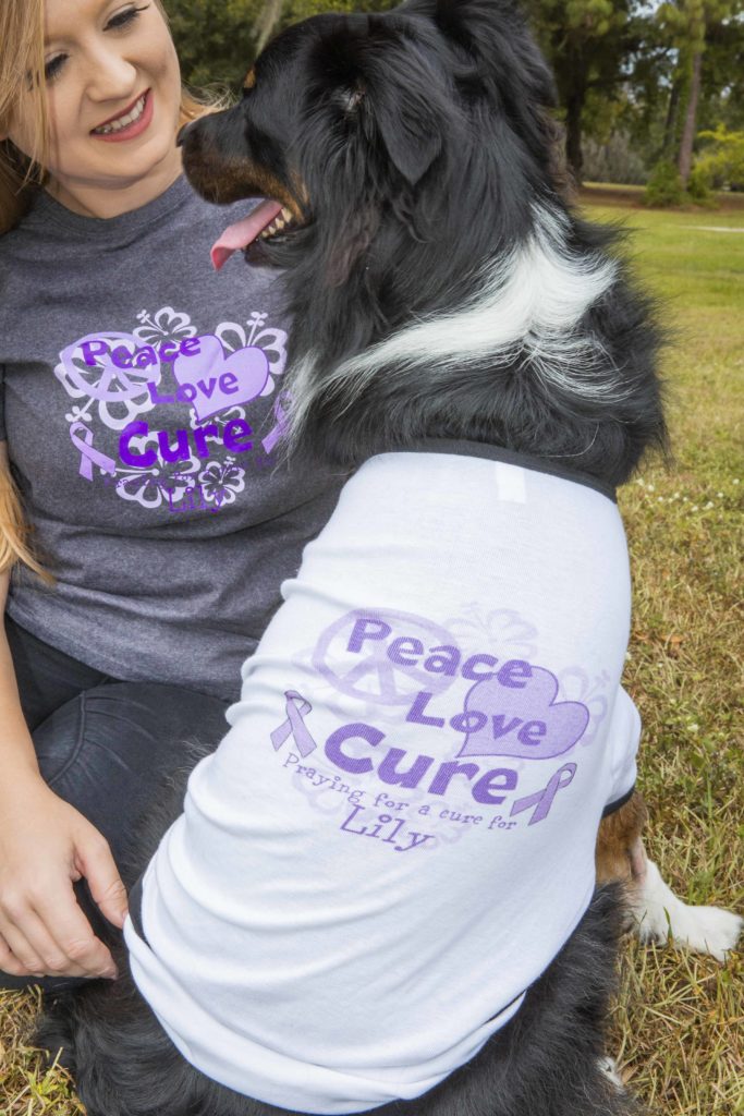 Girl and dog look at each other both wearing a shirt that says praying for a cure for lily cancer shirt